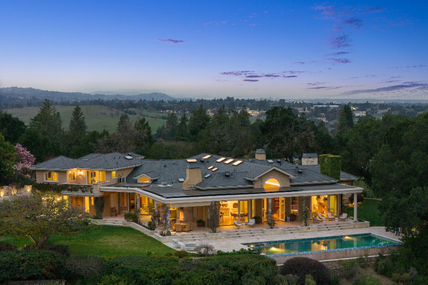 The funded Portola Valley estate at dusk: a broad residence with covered terraces, a lawn and a pool against wooded hills.